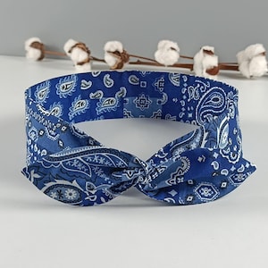 Può includere: Una fascia per capelli blu con un motivo paisley bianco e un dettaglio centrale annodato. Il tessuto presenta un design complesso e ripetitivo composto da motivi a goccia e forme geometriche su tutta la superficie dell'accessorio.