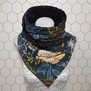 Puede incluir: Bufanda tipo cowl de color verde azulado oscuro con un estampado floral en blanco, amarillo y azul. La bufanda tiene un forro negro texturizado y se abrocha con un broche plateado. El diseño presenta flores y hojas estilizadas.