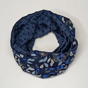 Pode incluir: Um cachecol infinito azul marinho com um padrão de folhas em branco, azul e dourado. O interior é um tecido azul escuro macio com uma textura pontilhada. Este cachecol é ideal para o inverno.