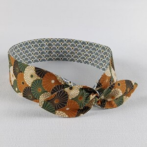 Puede incluir: Una diadema de tela con un diseño anudado. La diadema presenta un estampado floral en tonos marrón, verde y blanco sobre un fondo crema. El forro interior tiene un patrón geométrico en forma de abanico en gris, blanco y amarillo.