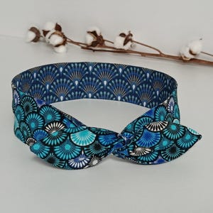 Può includere: Una fascia blu con un fiocco annodato. Il tessuto presenta un motivo ripetuto di forme a ventaglio turchese, blu e bianco. Un accessorio per capelli alla moda.