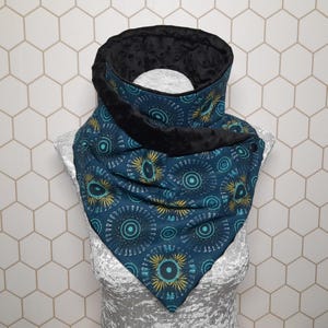 May include: A teal and black neck warmer with a geometric pattern. The neck warmer has a black, plush lining and a button closure. The pattern features teal, yellow, and white circular designs on a dark teal background. The neck warmer is displayed on a mannequin.