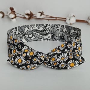 Op de afbeelding: Een zwart-witte bloemen haarband met een geknoopt ontwerp. De haarband heeft een madeliefjespatroon aan de voorkant en een botanische print aan de binnenkant. De madeliefjes hebben gele centra.