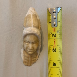 May include: A carved stone sculpture of a person's head with a headdress. The sculpture is approximately 3 inches tall and is made of a light brown stone.
