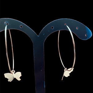 May include: A pair of silver hoop earrings with butterfly charms hanging from them. The earrings are displayed on a black earring stand.