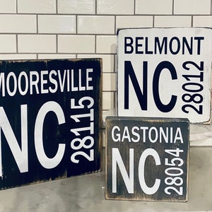 May include: Three wooden signs with black and white lettering. The signs read "Mooresville NC 28115", "Belmont NC 28012", and "Gastonia NC 28054".