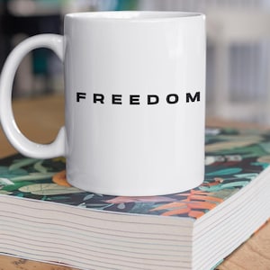 May include: A white ceramic mug with the word "FREEDOM" printed in black. The mug is sitting on top of a book with a colorful floral design. The background is blurred, showing a wooden table and a window.