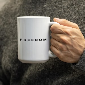 May include: White ceramic mug with the word "FREEDOM" printed in black. The mug has a standard handle and is held by a person wearing a gray sweater. The mug is a standard size for coffee or tea.