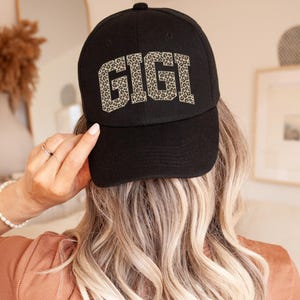 May include: Black baseball cap with the word "GIGI" in leopard print lettering. The cap is a classic six-panel design with a curved brim. The hat is made of a soft, durable fabric.