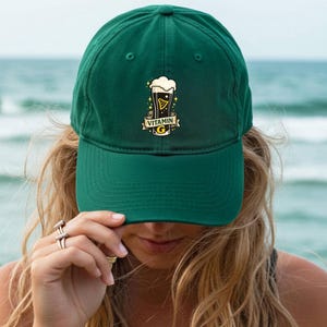 May include: A dark green baseball cap with a curved brim. The cap features a graphic of a beer glass with the text "Vitamin G". The background is a blurred view of the ocean.