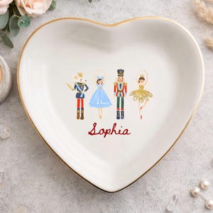 May include: A white heart-shaped ceramic trinket dish with a gold-painted rim. The center features a colorful illustration of a mouse soldier, a ballerina in a blue dress, a nutcracker, and a ballerina in a gold dress, with the name Sophia in red.