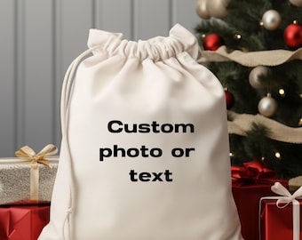 May include: A cream-colored drawstring gift bag with the text "Custom photo or text" in black. The bag is in front of a decorated Christmas tree and wrapped gifts.
