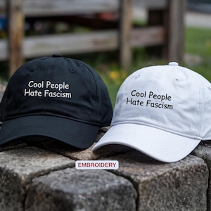 May include: Two baseball caps, one black and one white, are displayed. Each cap has the embroidered text "Cool People Hate Fascism" in white. The caps are resting on a stone surface.