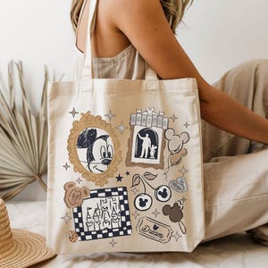 Puede incluir: Bolso tote de lona beige con un collage tem&aacute;tico de Disney. El dise&ntilde;o incluye un boceto de Mickey Mouse, un castillo, cerezas y la palabra "Dream". El bolso tiene dos asas.