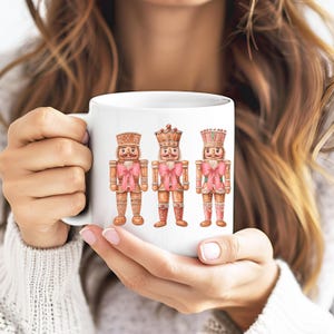 Puede incluir: Una taza de cerámica blanca con una ilustración impresa de tres figuras de cascanueces. Cada cascanueces está decorado en tonos marrones y rosas, luciendo un lazo rosa a juego en el cuello. Las figuras están dispuestas una al lado de la otra.