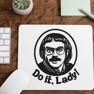 May include: White mousepad with a black and white illustration of a man with glasses and a mustache inside a circle. The text "Do it, Lady!" is below the image. A keyboard, mouse, pen, and sticky notes are on a wooden desk.