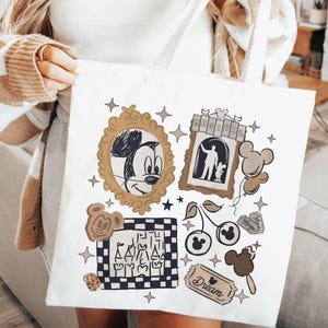 Puede incluir: Bolso tote blanco con ilustraciones tem&aacute;ticas de Disney. Incluye un boceto de Mickey Mouse en un marco dorado, una silueta de padre e hijo y otros gr&aacute;ficos. La palabra "Magical" es visible.