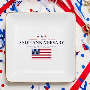 Peut inclure: Un plateau carré en céramique blanche avec un bord doré. Le plateau porte l'inscription "UNITED STATES OF AMERICA 250TH ANNIVERSARY 1776-2026" et un petit drapeau américain. Des confettis et des serpentins rouges, blancs et bleus entourent le plateau.
