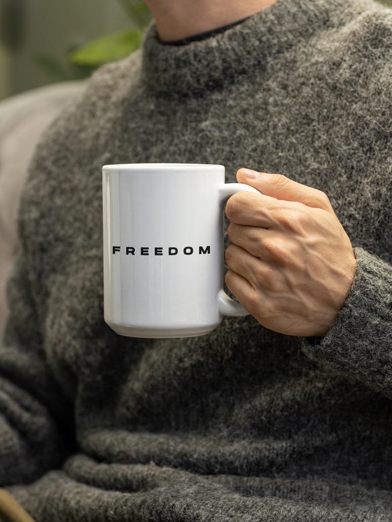 May include: White ceramic mug with the word "FREEDOM" printed in black. The mug has a standard handle and is held by a person wearing a gray sweater. The mug is a standard size for coffee or tea.