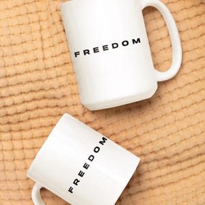May include: Two white ceramic mugs with the word "FREEDOM" printed in black. The mugs have a simple design with a curved handle. The mugs are on a textured, tan-colored surface.