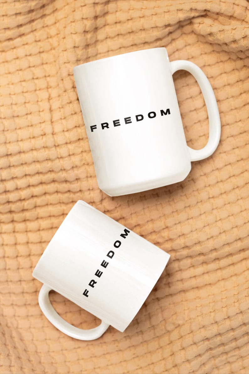 May include: Two white ceramic mugs with the word "FREEDOM" printed in black. The mugs have a simple design with a curved handle. The mugs are on a textured, tan-colored surface.