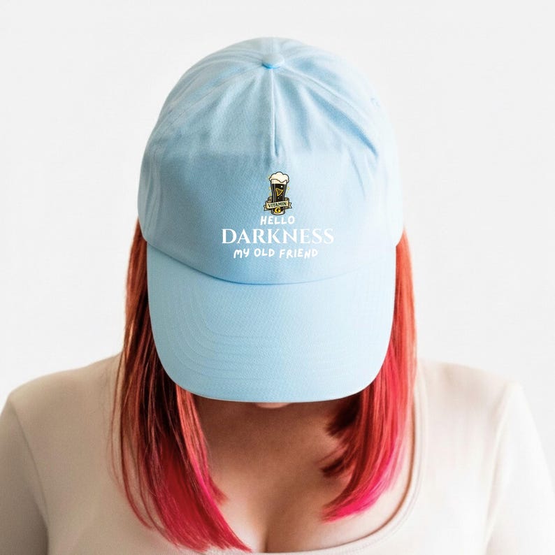 May include: Light blue baseball cap with the text "HELLO DARKNESS MY OLD FRIEND" in white. The cap features a small beer graphic with the word "VITAMIN B" above it. The cap is made of a soft material.