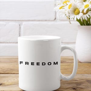 May include: White ceramic mug with the word "FREEDOM" printed in black. The mug is on a wooden surface, with a white vase of daisies in the background. The mug has a curved handle.