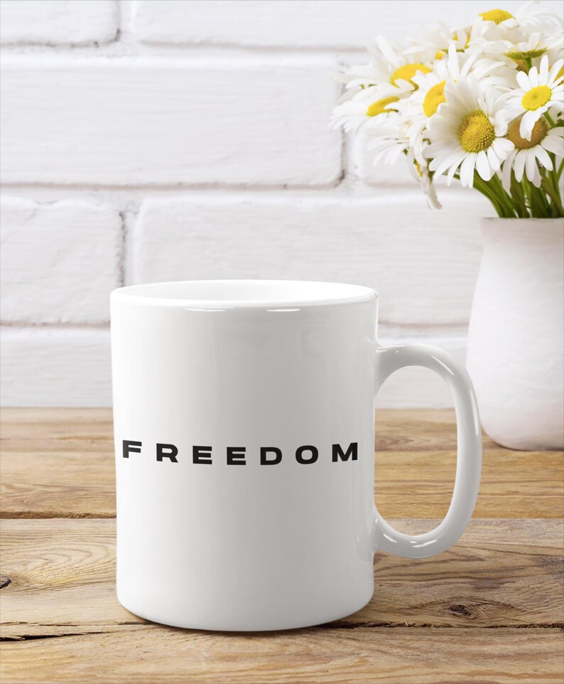 May include: White ceramic mug with the word "FREEDOM" printed in black. The mug is on a wooden surface, with a white vase of daisies in the background. The mug has a curved handle.