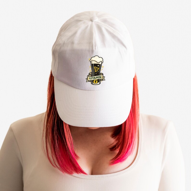 May include: White baseball cap featuring a graphic of a pint of dark beer with a foamy head. The design includes the text "Vitamin G" on a banner. The cap is a classic six-panel design with a curved brim.