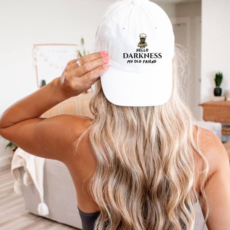 May include: A white baseball cap with the text "HELLO DARKNESS MY OLD FRIEND" in black lettering. The cap features a small graphic of a beer mug. The hat is being held by a person with long blonde hair.