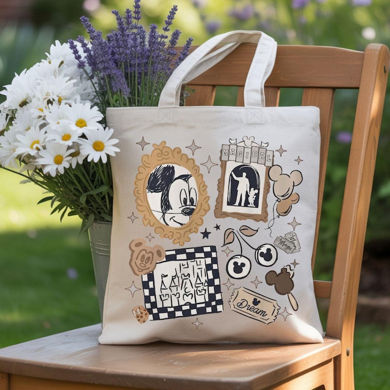 Puede incluir: Bolso tote beige con ilustraciones de Disney en marr&oacute;n y negro, incluyendo a Mickey Mouse. El bolso tiene asas color crema y est&aacute; sobre una silla de madera junto a un ramo de margaritas blancas y lavanda morada. La palabra "Dream" es visible.