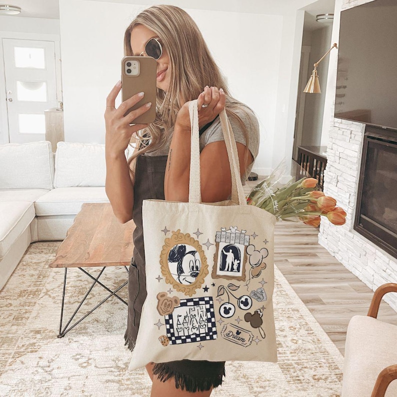 Puede incluir: Bolso tote de color crema con ilustraciones tem&aacute;ticas de Disney, incluyendo Mickey Mouse en un marco dorado. El bolso tiene forma rectangular con dos asas. Otras im&aacute;genes incluyen un patr&oacute;n de cuadros blanco y negro y la palabra "Dream".