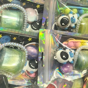May include: A collection of packaged space-themed items. The packages contain astronaut keychains, colorful balls, and small containers with glittery substances. The packaging is clear with iridescent accents, creating a playful and eye-catching display.