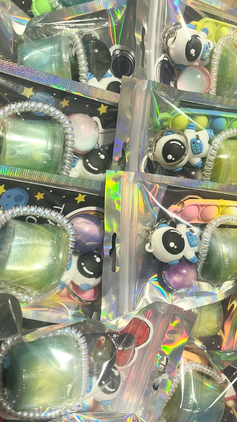 May include: A collection of packaged space-themed items. The packages contain astronaut keychains, colorful balls, and small containers with glittery substances. The packaging is clear with iridescent accents, creating a playful and eye-catching display.
