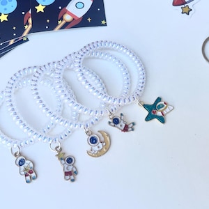 May include: White spiral hair ties with space-themed charms, including astronauts, a star, and a crescent moon. A colorful pop-it toy and a space-themed sticker sheet are also visible. The charms are small and detailed.