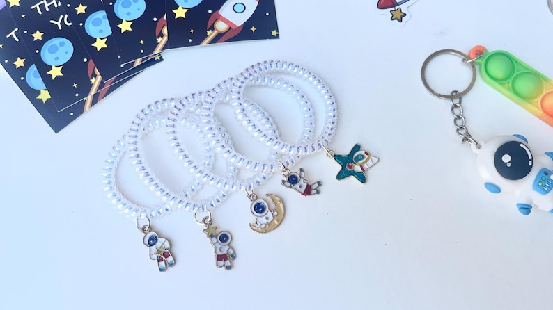 May include: White spiral hair ties with space-themed charms, including astronauts, a star, and a crescent moon. A colorful pop-it toy and a space-themed sticker sheet are also visible. The charms are small and detailed.
