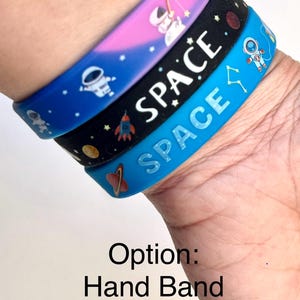 May include: Three colorful space-themed wristbands stacked on top of each other. The top band is pink and purple, the middle band is black, and the bottom band is blue. Each band features white text that says "SPACE" and space-related graphics.