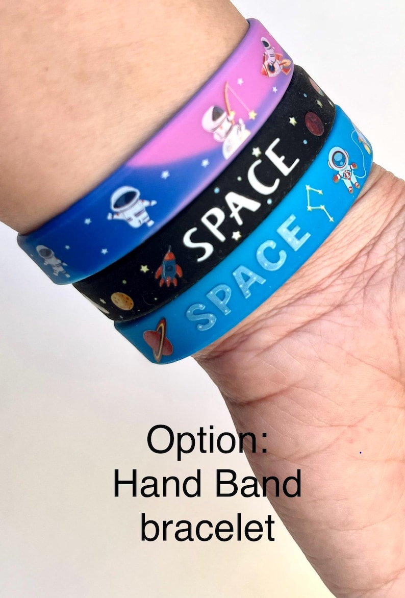 May include: Three colorful space-themed wristbands stacked on top of each other. The top band is pink and purple, the middle band is black, and the bottom band is blue. Each band features white text that says "SPACE" and space-related graphics.