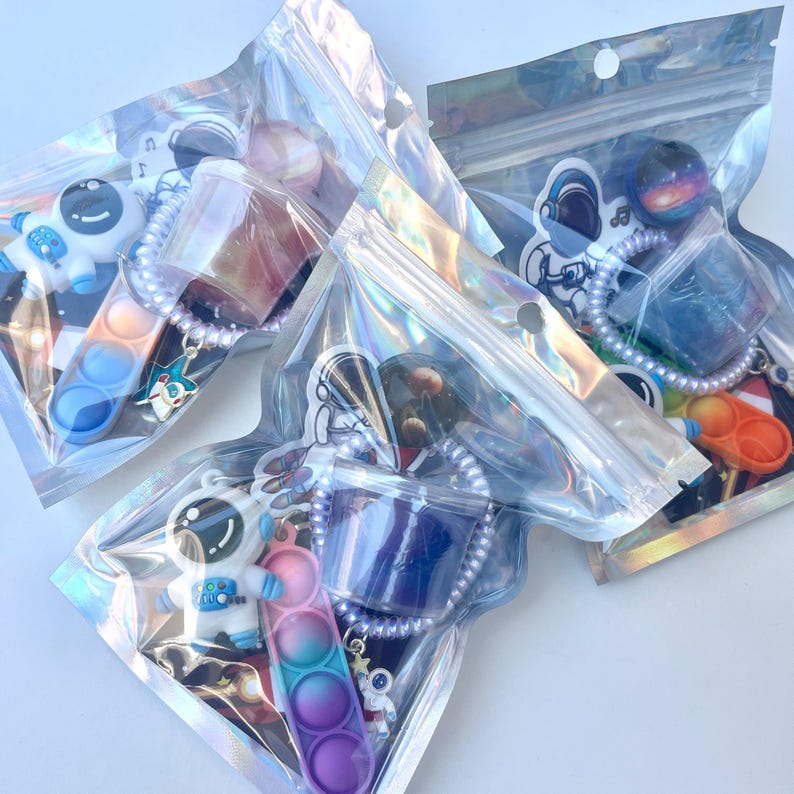 May include: Several clear plastic bags containing astronaut-themed keychains and fidget toys. The keychains feature astronaut figures, space-themed charms, and colorful pop-it toys. The bags are sealed and ready for sale.