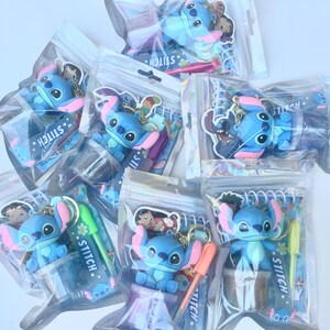 May include: A collection of Stitch character keychains and pens in clear plastic bags. The keychains feature a blue alien with large ears and a pink nose. Some bags include pens with the word "Stitch" printed on them.