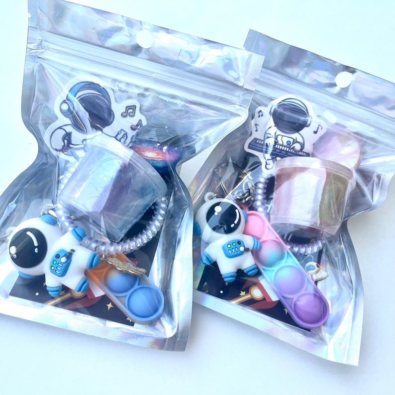 May include: Two clear plastic bags each containing a keychain with an astronaut figure, a small container, and a colorful pop-it toy. The astronaut keychains are white and blue. The pop-it toys have blue, pink, and purple bubbles.