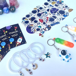 May include: An assortment of space-themed items, including keychains, stickers, bracelets, and thank you cards. The keychains feature astronaut figures and colorful pop-it toys. The cards read "THANK YOU!" with rocket and planet illustrations.