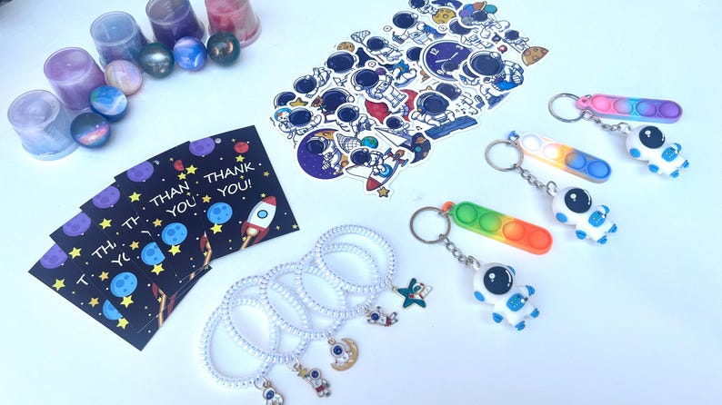 May include: An assortment of space-themed items, including keychains, stickers, bracelets, and thank you cards. The keychains feature astronaut figures and colorful pop-it toys. The cards read "THANK YOU!" with rocket and planet illustrations.