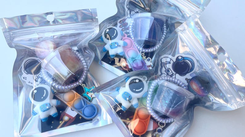 May include: Several clear, iridescent bags filled with space-themed keychains and toys. Each bag contains a small astronaut figure, a pop-it toy, a rocket ship, and a small container of slime. The keychains are attached to a beaded chain.