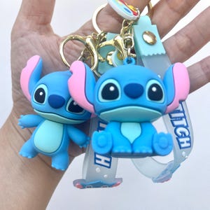 May include: Two blue Stitch keychains with pink ears and large black eyes. One keychain is standing, the other is sitting. The keychains are attached to gold-colored metal rings and a light blue strap with the word "Stitch".