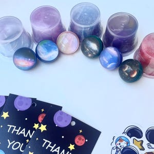 May include: An assortment of space-themed items, including colorful planet-shaped plugs, clear containers, thank you cards with planets and stars, and astronaut stickers. The plugs are in shades of blue, purple, and pink.