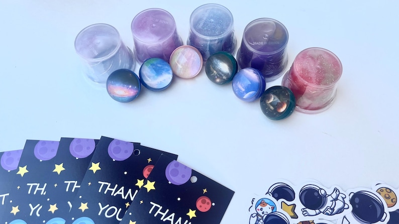 May include: An assortment of space-themed items, including colorful planet-shaped plugs, clear containers, thank you cards with planets and stars, and astronaut stickers. The plugs are in shades of blue, purple, and pink.