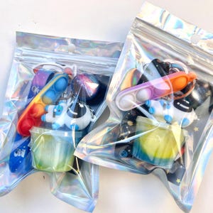 May include: Two iridescent zipper bags filled with space-themed toys. Each bag contains a colorful pop-it, astronaut keychain, a small container of slime, and other space-related trinkets. The bags are sealed and ready for gifting.