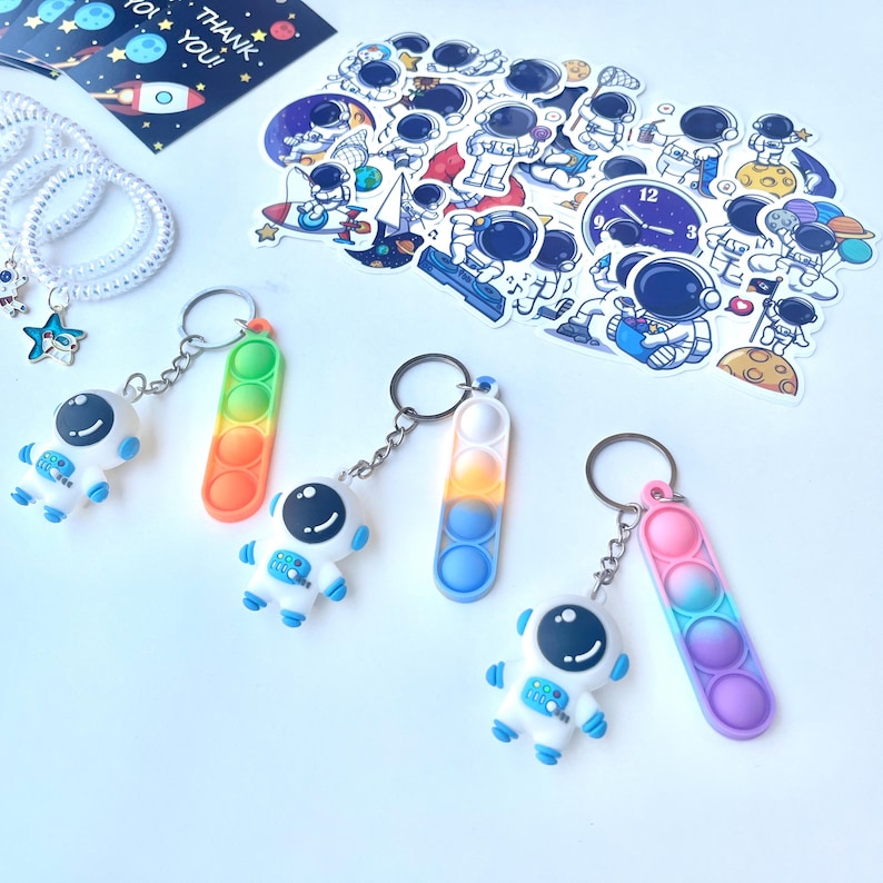 May include: A collection of space-themed items, including astronaut keychains with colorful bubble pop toys, stickers, and a bracelet with star and astronaut charms. The keychains feature white astronaut figures with blue accents and a variety of rainbow-colored bubble pop toys.