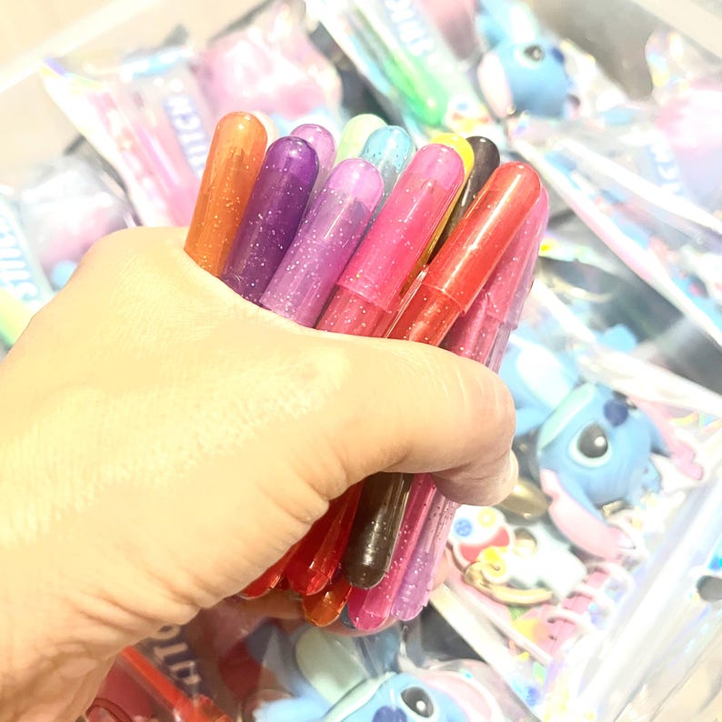 May include: A hand holding a bundle of colorful glitter glue pens. The pens are various shades of red, pink, purple, orange, and brown. The pens are cylindrical and have glitter inside. Other items are visible in the background.